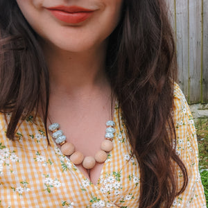 A mum wearing a teething and breastfeeding necklace made of beech and leaf print silicone beads with rose gold spacers threaded onto a waxed cord. She is wearing a yellow gingham and daisy print dress and is sat on the grass. 