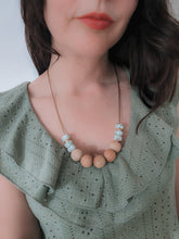 Load image into Gallery viewer, A woman wearing a sage green leaf print and beech beaded teething fiddle necklace with rose gold spacers, threaded on a wax cord. She is wearing the necklace with a sage green top. 
