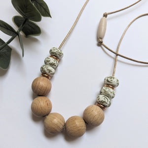 Teething and breastfeeding necklace with beech and leaf print silicone beads and rose gold spacers threaded on a waxed cord on a white background next to a eucalyptus leaf