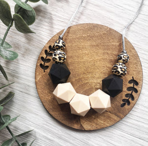 Leopard print, black and nude silicone teething and breastfeeding necklace on a soft grey cord, lying on a wooden disc with a eucalyptus stem to the side.